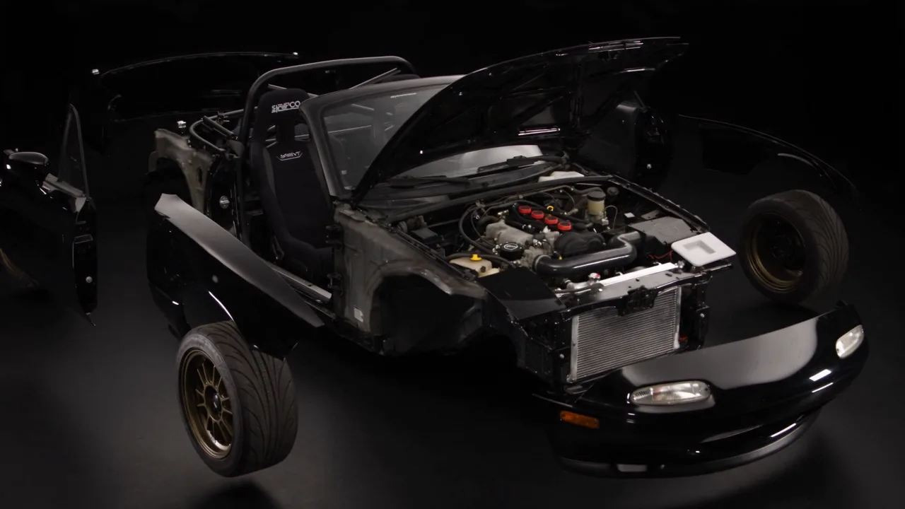 A 1994 Mazda MX-5 separated into its panels, wheels and engine, held apart.